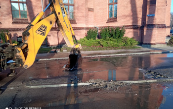 To zdjęcie przedstawia żółtą koparkę JCB z opuszczoną łyżką, stojącą na mokrym asfalcie, gdzie widoczny jest rozkopany fragment nawierzchni z kałużami wody.