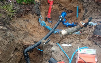 Zdjęcie przedstawia wykop ziemny, w którym trwa montaż. Widoczne są rury: czerwony hydrant nadziemny i niebieskie elementy armatury, w tym zasuwy kołnierzowe, połączone z czarnymi rurami.