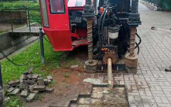 To zdjęcie przedstawia czerwoną wiertnicę do wykonywania bezwykopowego przewiertu, który umożliwi instalację nowego wodociągu. Maszyna, stoi na kostce brukowej, z osprzętem.