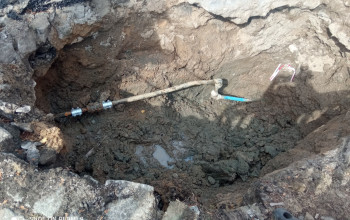 Na zdjęciu widoczny jest odcinek rury, który biegnie w wykopie ziemnym, częściowo zanurzony w błocie. Rura ma na sobie duży metalowy zacisk naprawczy.