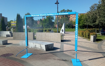 Zdjęcie przedstawia niebieską, metalową ramę w kształcie bramy, pełniącą funkcję kurtyny wodnej, z dysz tryska drobna mgiełka. Instalacja znajduje się na placu wyłożonym kostką brukową.