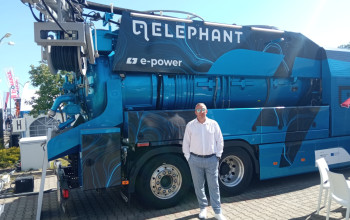 Duży, niebieski samochód ciężarowy do czyszczenia kanalizacji stoi na placu wyłożonym kostką brukową. Obok pojazdu stoi mężczyzna w jasnej koszuli i spodniach.