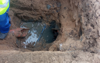 Wykop ziemny z awarią na przyłączu wodociągowym, wypełniony błotem i wodą.
Widoczny jest fragment czerwonej rury.